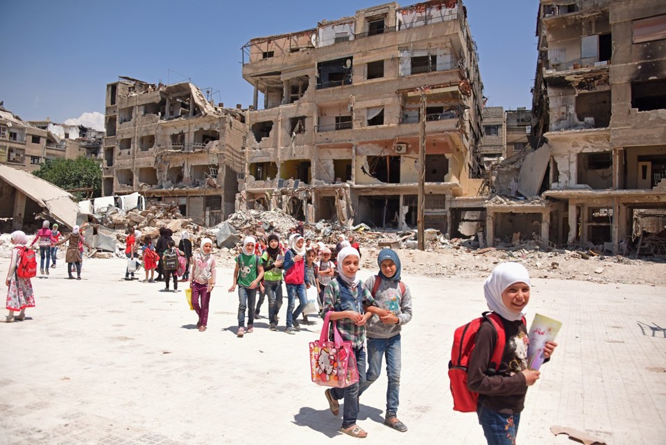 Skolejenter i Syria går sammen med ruiner i bakgrunnen.