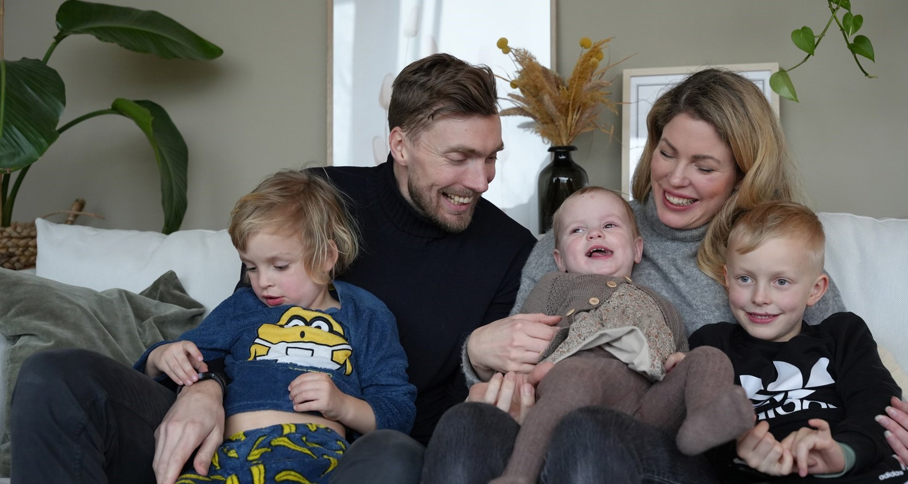 En familie med to voksne og tre barn smiler og sitter i en sofa.